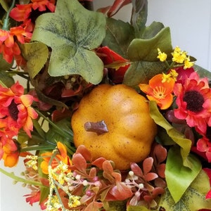 May include: A fall-themed wreath with red, orange, and yellow flowers, green leaves, and a small brown pumpkin.