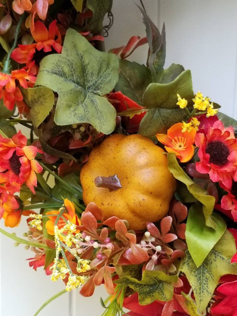 May include: A fall-themed wreath with red, orange, and yellow flowers, green leaves, and a small brown pumpkin.
