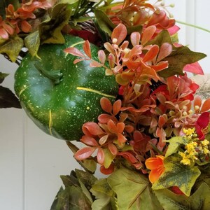 May include: A green gourd with a speckled surface is nestled in a wreath of fall foliage. The wreath features red, orange, and yellow leaves, as well as small white flowers.
