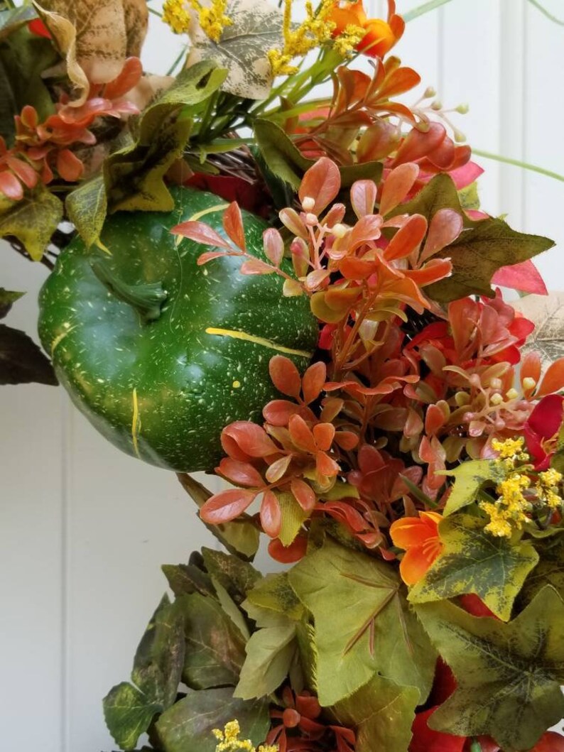 May include: A green gourd with a speckled surface is nestled in a wreath of fall foliage. The wreath features red, orange, and yellow leaves, as well as small white flowers.