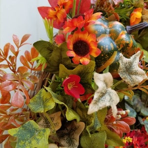 May include: A fall-themed wreath with a green, orange, and brown gourd, red and orange flowers, and green and brown leaves.