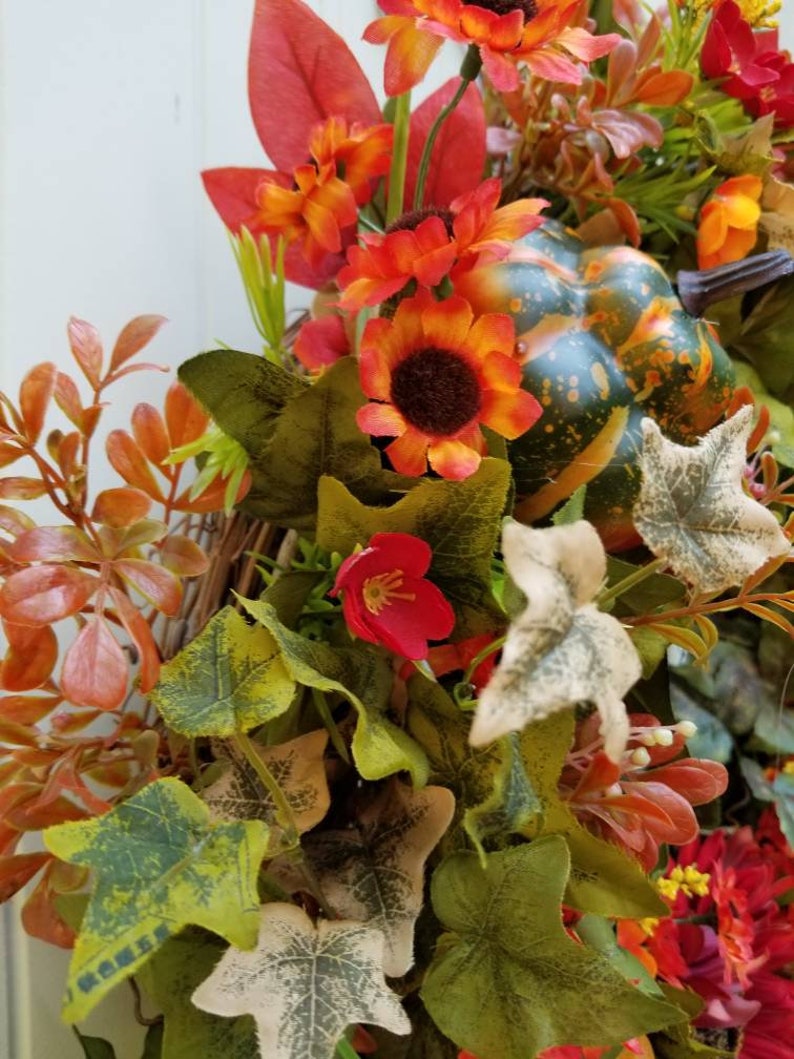 May include: A fall-themed wreath with a green, orange, and brown gourd, red and orange flowers, and green and brown leaves.