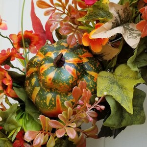 May include: A fall-themed wreath with a green, orange, and yellow gourd, red and orange flowers, and green leaves.