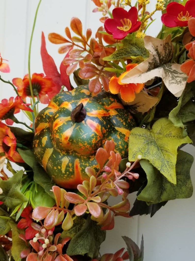 May include: A fall-themed wreath with a green, orange, and yellow gourd, red and orange flowers, and green leaves.