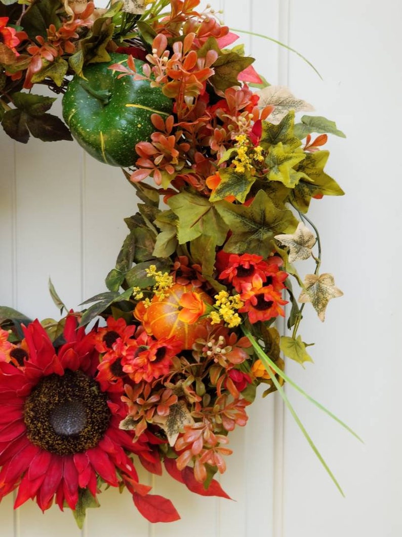 May include: A fall-themed wreath with red sunflowers, orange gourds, and green and brown leaves.