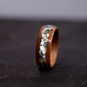 May include: A wooden ring with a band of iridescent abalone shell inlay. The ring is dark brown and the shell inlay is a mix of white, blue, and green.
