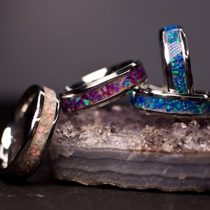 May include: Four silver rings with different colored opal inlays. The rings are stacked on top of each other and are sitting on a piece of amethyst.