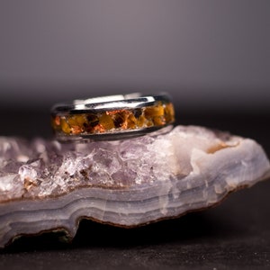 May include: A silver ring with a band of orange and brown stones set in a silver setting. The ring is resting on a purple and white crystal.