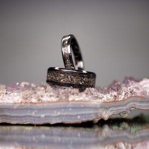 May include: Two dark metal rings featuring a textured, granular inlay around the center of the bands. The rings are positioned upright, resting on a horizontal slice of purple and white amethyst crystal with visible layered banding and jagged edges.
