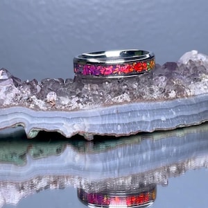 May include: A silver ring with a colorful, mosaic-like inlay of pink, red, and orange hues. The ring is set against a backdrop of a crystal formation, with its reflection visible below. The ring's design is eye-catching and unique.