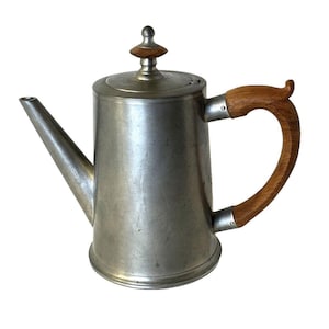 May include: A vintage metal coffee pot with a wooden handle and lid finial. The pot has a long spout and a curved handle. The metal has a brushed finish. The wooden handle and lid finial are a light brown color.