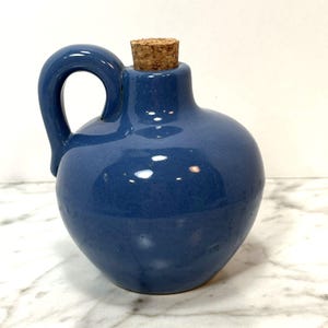 May include: A small, rounded ceramic jug with a glossy, solid blue glaze. The vessel features a single curved handle on the left side, a narrow neck, and a cylindrical cork stopper inserted into the top opening, creating a smooth, uniform finish.
