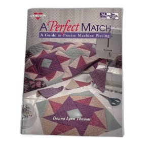 May include: A softcover instructional book titled A Perfect Match: A Guide to Precise Machine Piecing by Donna Lynn Thomas. The cover features a purple and beige star-patterned quilt, a sewing machine, fabric scraps, and a pair of scissors.