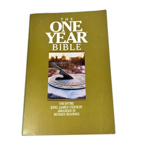 May include: A gold-colored book titled The One Year Bible with white text. The cover features a central rectangular photograph of a sundial on a stone base. Below the image, text reads The Entire King James Version Arranged in 365 Daily Readings.