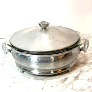 May include: A round, hammered silver-tone metal casserole dish with a clear glass insert. It features two side handles, a matching domed lid with a flower-shaped finial, and small feet. The metal surface has a textured, dimpled finish throughout.
