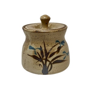 May include: A tan ceramic jar with a lid. The jar has a rounded shape with a slightly wider base and a narrower opening. It is decorated with brown and blue floral designs. The lid has a small knob for easy removal.