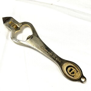 May include: A silver-toned bottle opener with a heart-shaped opening and a gold-colored oval end. The oval end features a gold-colored "CF" logo. The text "CALVERT DIST. CO., N.Y.C." is printed on the handle.