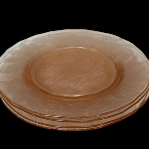 May include: A stack of four vintage pink glass plates. Each plate features a floral pattern around the rim and center. The plates are stacked on top of each other, showcasing the design and color.