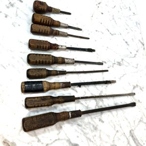May include: A collection of vintage screwdrivers with wooden handles and metal shafts. The handles are various shades of brown, showing signs of wear. The metal shafts are dark, with flat or Phillips head tips. The screwdrivers are arranged on a white surface.