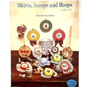 May include: A craft book titled Skirts, Scoops and Hoops by Joyce Seebo featuring thirty detailed designs. The cover displays various cross-stitch projects in embroidery hoops, fabric-covered jar lids, and a pitcher on a wooden surface.