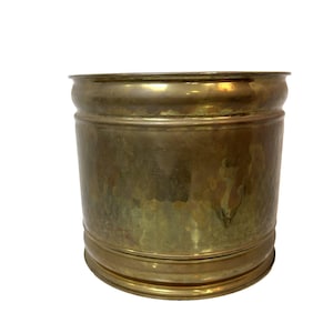 May include: A cylindrical brass planter with a hammered texture. The planter has a wide, rounded rim and a base with a similar rounded design. The brass has a warm, golden hue, and the surface shows signs of age.
