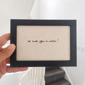 May include: A rectangular black picture frame containing a light beige fabric insert. The fabric features the embroidered black text "I love you a million!" written in a casual, handwritten script style centred within the frame.