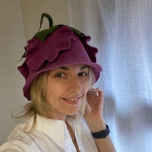 Puede incluir: Una mujer lleva un sombrero de fieltro morado con forma de flor con hojas verdes. El sombrero tiene un ala ancha y está decorado con bordes con volantes.
