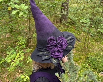 May include: A black and purple witch hat with a tall, pointed crown. The hat features a wide brim and is adorned with three purple felt roses and green leaves. The hat is a whimsical accessory.