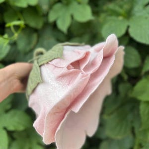 Pode incluir: Uma flor de feltro rosa claro feita &agrave; m&atilde;o, com um caule e folhas verdes. A flor tem m&uacute;ltiplas camadas de p&eacute;talas, criando uma apar&ecirc;ncia de rosa realista. O fundo &eacute; uma folhagem verde desfocada.