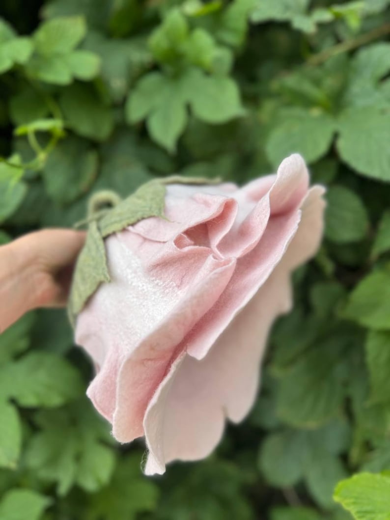 Pode incluir: Uma flor de feltro rosa claro feita &agrave; m&atilde;o, com um caule e folhas verdes. A flor tem m&uacute;ltiplas camadas de p&eacute;talas, criando uma apar&ecirc;ncia de rosa realista. O fundo &eacute; uma folhagem verde desfocada.