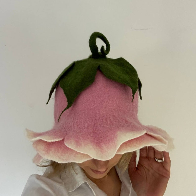 K&ouml;nnte beinhalten: Ein rosa Filzhut in Form einer Blume mit gr&uuml;nen Bl&auml;ttern und einem gekr&auml;uselten Stiel. Der Hut hat einen wei&szlig;en Rand an den Bl&uuml;tenbl&auml;ttern. Der Hut ist ein skurriles Accessoire.