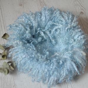 Puede incluir: Un accesorio de fotografía para recién nacidos en forma de nido azul claro y esponjoso. El nido está hecho de lana rizada suave y está rodeado por un anillo del mismo material. Unas hojas secas se colocan cerca del nido.
