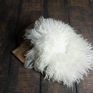 Puede incluir: Alfombra de piel de oveja blanca y esponjosa, perfecta para accesorios de fotograf&iacute;a de reci&eacute;n nacidos. La alfombra est&aacute; enrollada sobre un fondo de madera oscura.