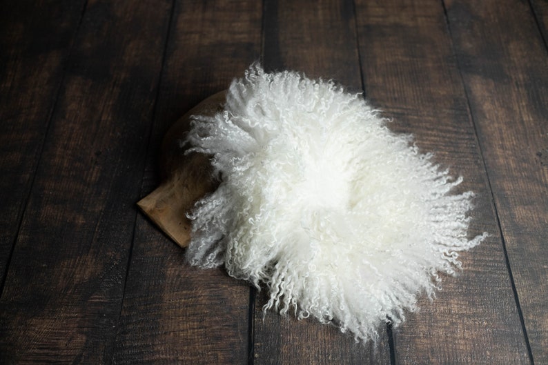 Puede incluir: Alfombra de piel de oveja blanca y esponjosa, perfecta para accesorios de fotograf&iacute;a de reci&eacute;n nacidos. La alfombra est&aacute; enrollada sobre un fondo de madera oscura.