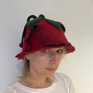 Puede incluir: Un sombrero de fieltro rojo con forma de rosa con hojas de fieltro verde. El sombrero lo lleva una persona con el pelo rubio.