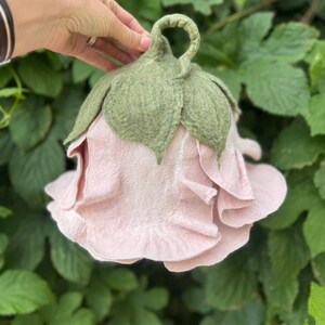 Pode incluir: Uma flor de feltro rosa claro feita &agrave; m&atilde;o, com folhas e um caule de feltro verde. A flor tem m&uacute;ltiplas camadas de p&eacute;talas e &eacute; segurada por uma m&atilde;o contra um fundo de folhagem verde. Um item decorativo &uacute;nico.