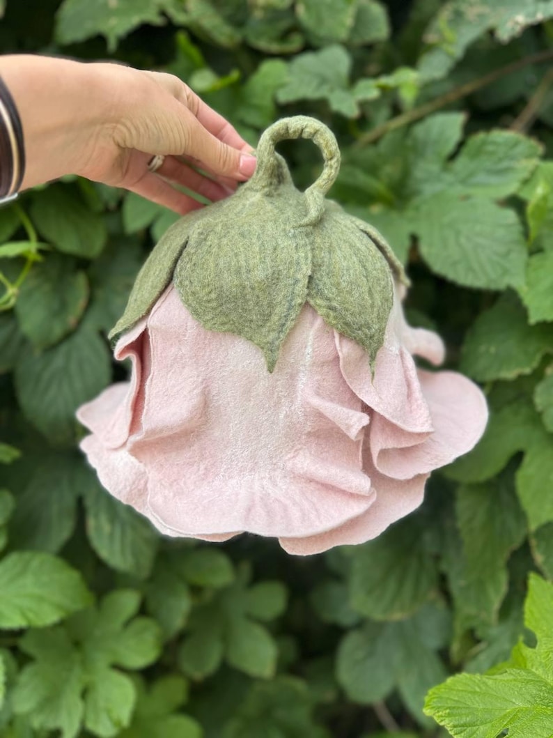 Pode incluir: Uma flor de feltro rosa claro feita &agrave; m&atilde;o, com folhas e um caule de feltro verde. A flor tem m&uacute;ltiplas camadas de p&eacute;talas e &eacute; segurada por uma m&atilde;o contra um fundo de folhagem verde. Um item decorativo &uacute;nico.