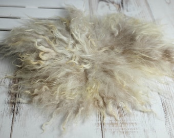 May include: A light beige, curly sheepskin rug. The rug has a soft, fluffy texture and is placed on a white wooden surface. The sheepskin has a natural, organic look, with variations in color and curl.