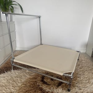Puede incluir: Una moderna y minimalista cama para perros con estructura cromada y cama de lona beige. La cama tiene forma rectangular y está elevada del suelo. La cama está colocada sobre una alfombra marrón y peluda.