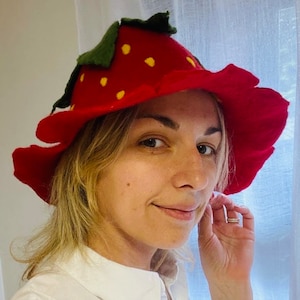Puede incluir: Un sombrero de fieltro rojo con forma de fresa con hojas verdes y semillas amarillas. El sombrero lo lleva una persona con el pelo rubio.
