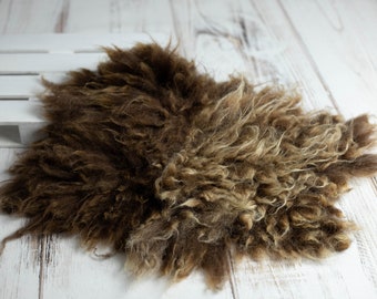 May include: A brown, fluffy sheepskin rug with a natural, textured appearance. The rug is irregularly shaped and lies on a white wooden surface. The fur has varying shades of brown and tan.