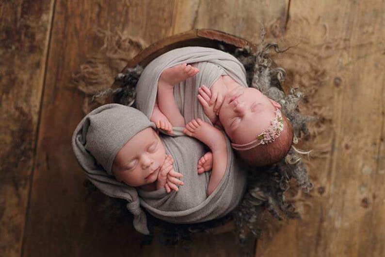 Puede incluir: Dos beb&eacute;s reci&eacute;n nacidos envueltos en tela gris, durmiendo pl&aacute;cidamente en un cuenco de madera. Un beb&eacute; lleva un gorro de punto gris, mientras que el otro lleva una diadema rosa con adornos florales.