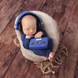 May include: A baby wearing blue headphones and a blue onesie is holding a blue toy boombox. The baby is sitting in a white furry chair on a wooden floor.