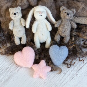 Pu&ograve; includere: Tre giocattoli di animali in feltro, un orso marrone, un coniglietto bianco e un orso marrone, con due cuori in feltro, uno rosa e uno blu, e una stella in feltro rosa su una superficie di legno bianca.