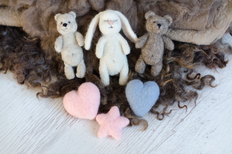 Pu&ograve; includere: Tre giocattoli di animali in feltro, un orso marrone, un coniglietto bianco e un orso marrone, con due cuori in feltro, uno rosa e uno blu, e una stella in feltro rosa su una superficie di legno bianca.