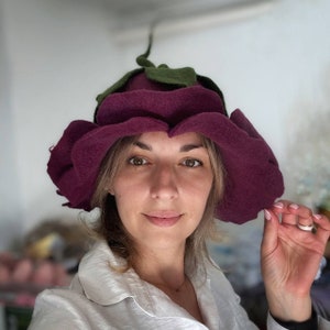 Puede incluir: Una mujer lleva un sombrero de fieltro morado con forma de flor con hojas de fieltro verde. El sombrero tiene una ala ancha y está decorado con múltiples capas de pétalos de fieltro.