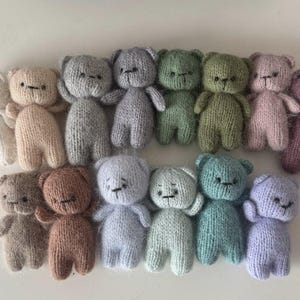 Op de afbeelding: Een verzameling van 15 gebreide teddyberen in verschillende kleuren, waaronder wit, beige, grijs, groen, roze, paars en blauw. De beren zijn allemaal klein en hebben zwarte ogen en neuzen.