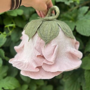 Pode incluir: Uma chaleira de ch&aacute; de feltro rosa claro feita &agrave; m&atilde;o em forma de rosa, com folhas de feltro verde e uma al&ccedil;a de la&ccedil;o. A chaleira &eacute; segurada contra um fundo de folhagem verde.