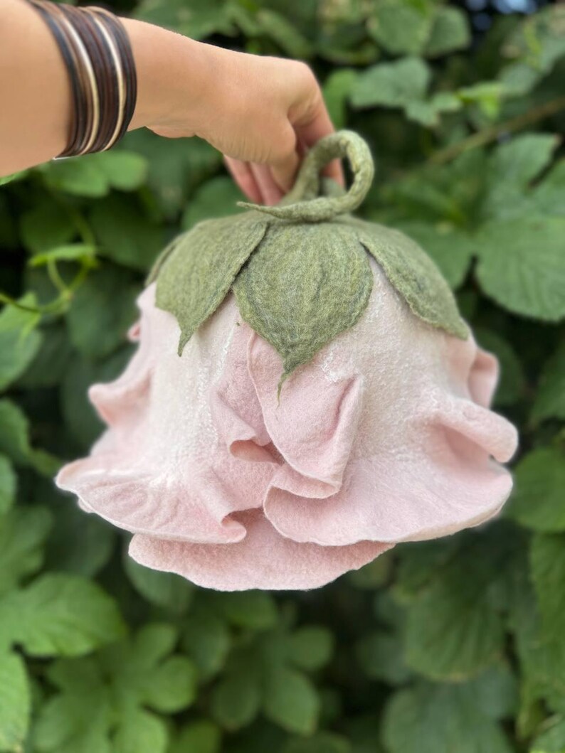 Pode incluir: Uma chaleira de ch&aacute; de feltro rosa claro feita &agrave; m&atilde;o em forma de rosa, com folhas de feltro verde e uma al&ccedil;a de la&ccedil;o. A chaleira &eacute; segurada contra um fundo de folhagem verde.