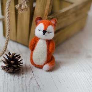 Puede incluir: Una pequeña figura de zorro de fieltro naranja y blanco con los ojos cerrados. El zorro está sentado sobre una superficie de madera blanca.